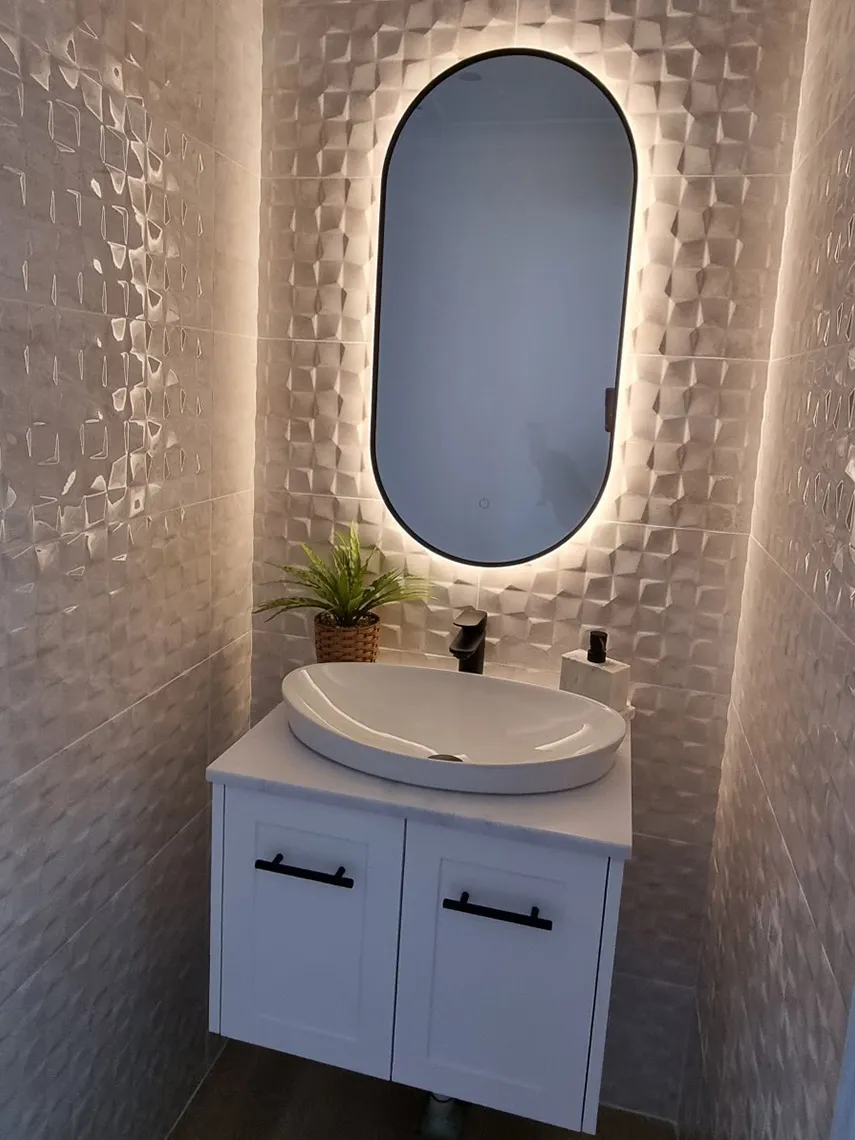 Backlit oval mirror above a white wall-hung vanity with a vessel basin and textured tiles