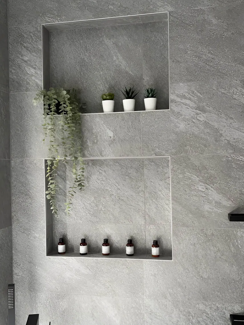 Two recessed shower niches in grey stone-look tiles holding small plants and bottles