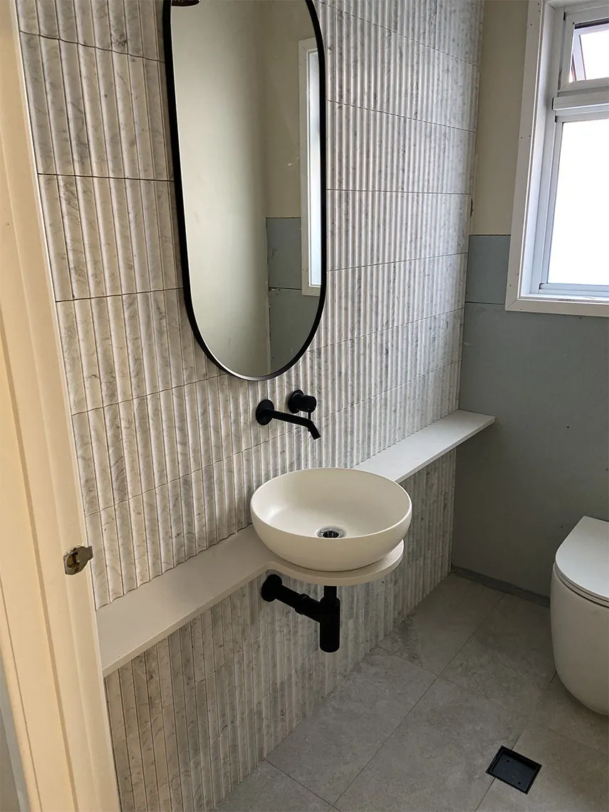 Round basin on a floating shelf below a black-framed oval mirror, with fluted wall tiles